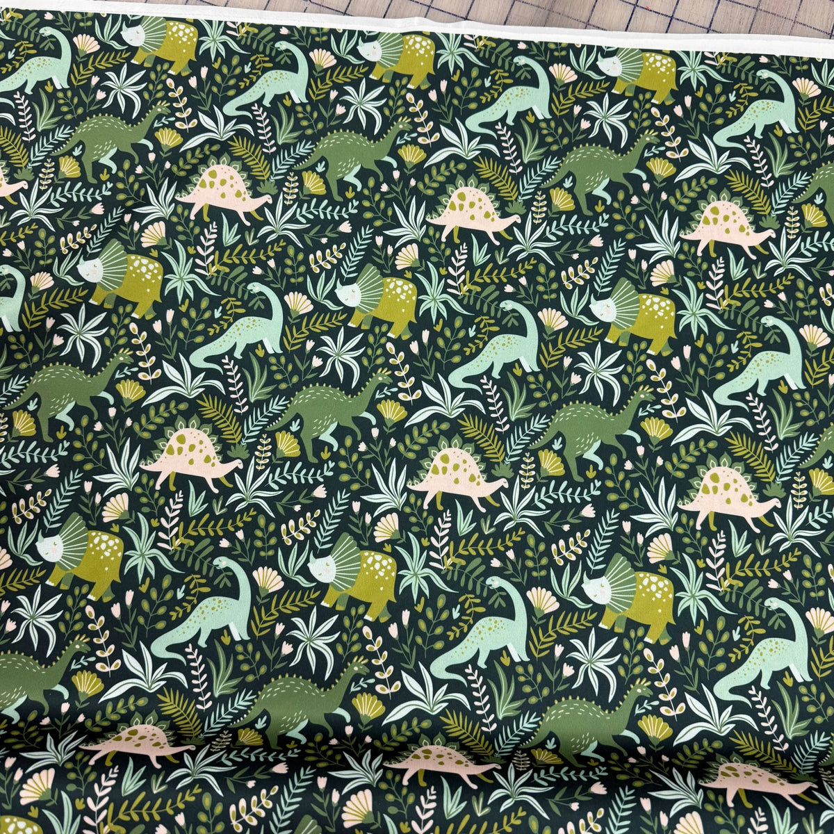 Dinosaur print fabric by Nature's Fabrics, featuring green and pink dinosaurs among floral patterns on dark background