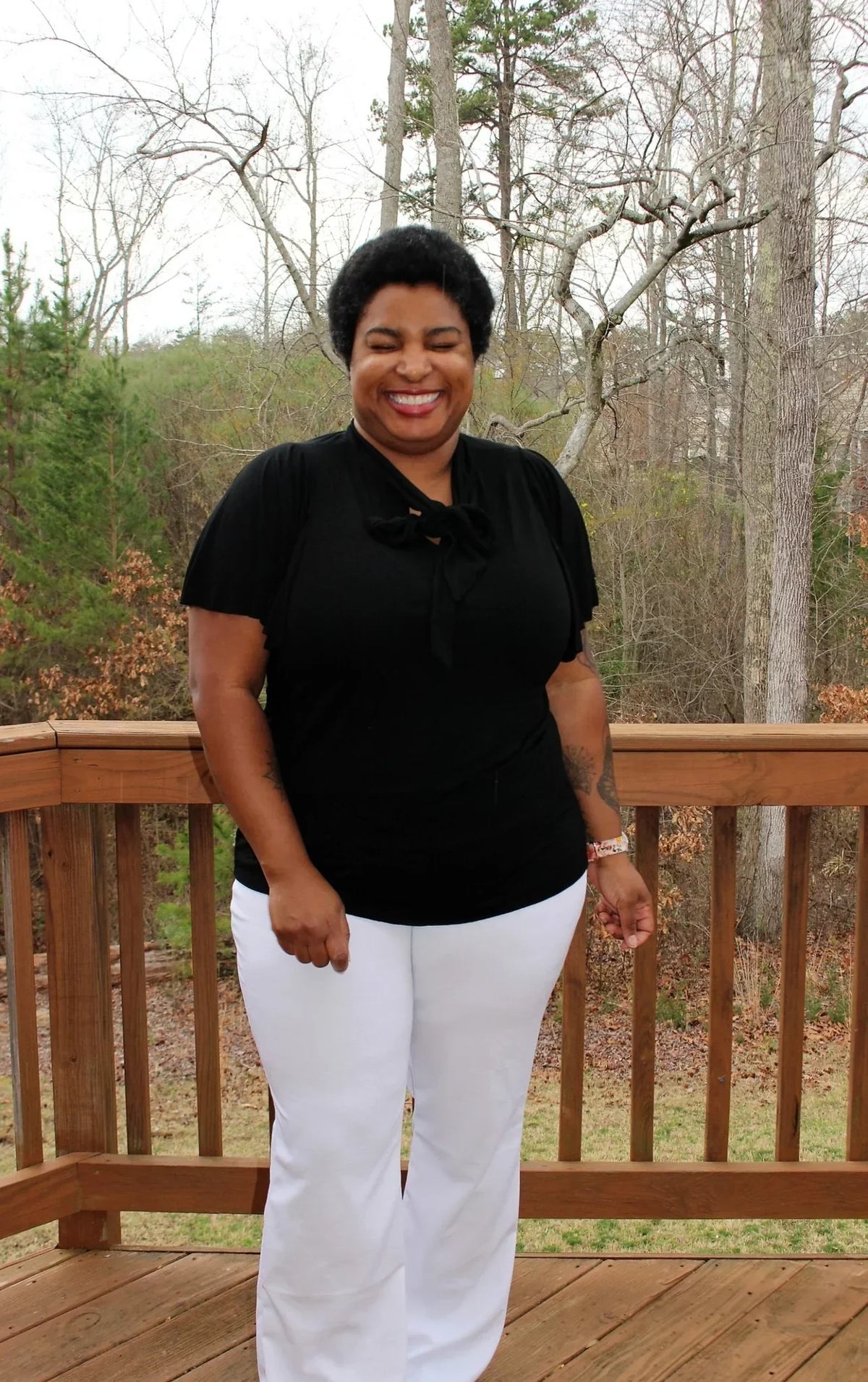A smiling woman wearing a Nature's Fabrics black blouse and white pants on a wooden deck.
