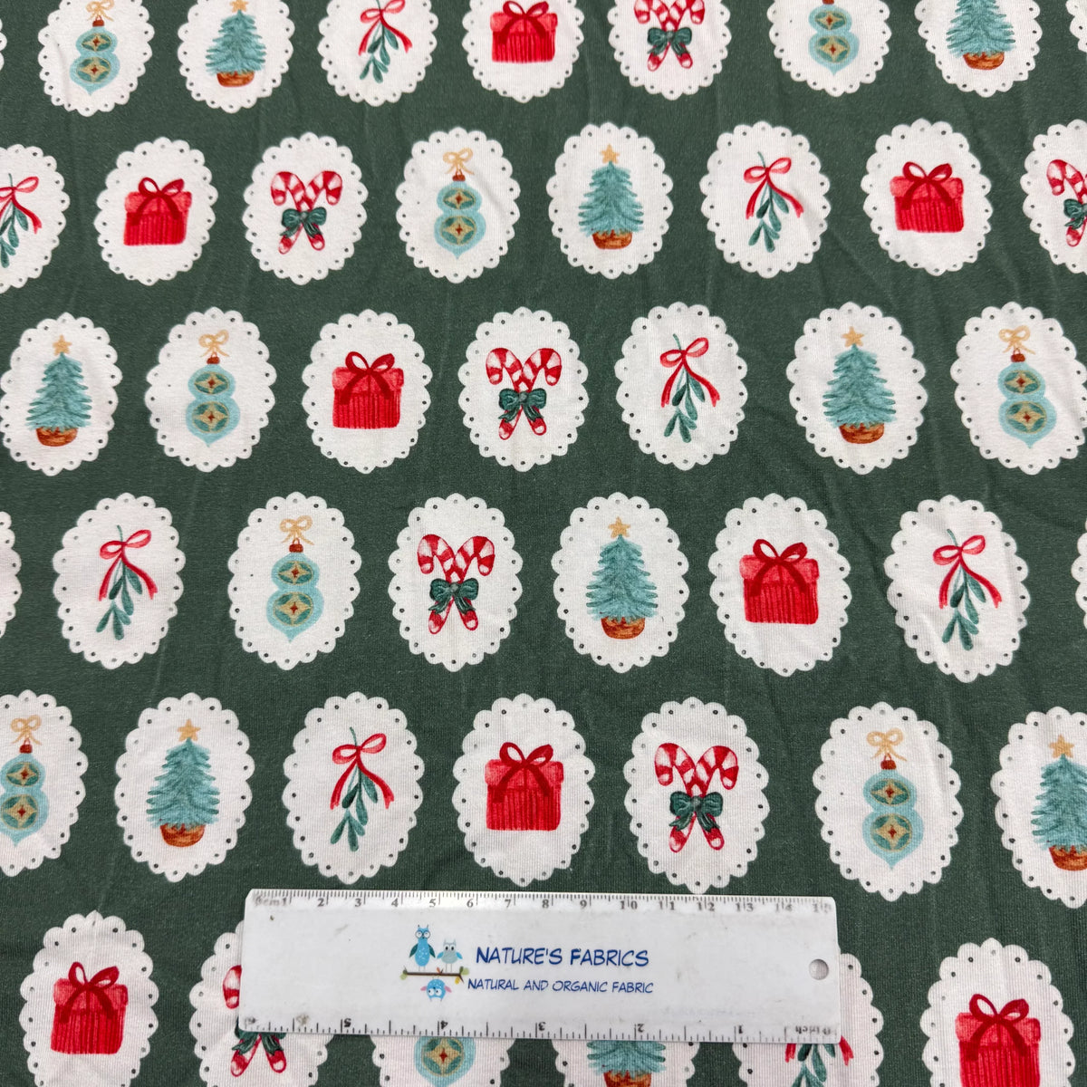 Nature's Fabrics Christmas-themed fabric with festive icons on green background, featuring trees, gifts, and candy canes in white doilies.