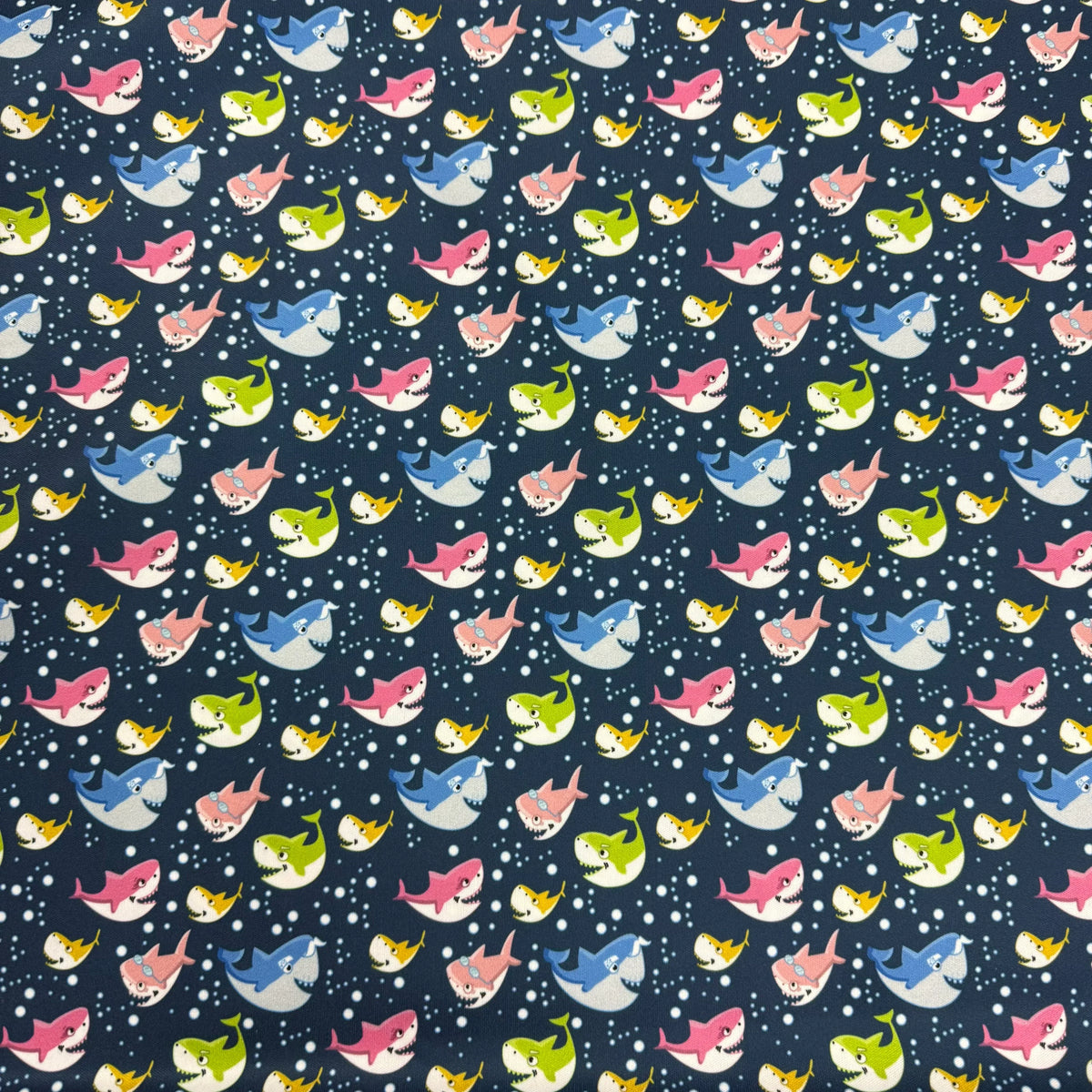 Colorful cartoon shark print fabric by Nature's Fabrics, featuring pink, blue, green, and yellow sharks on a navy background with white bubbles.