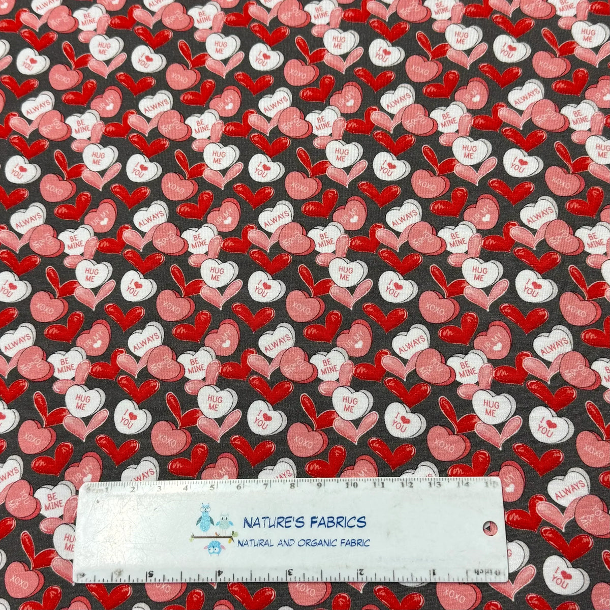 Valentine's Day heart candy print fabric by Nature's Fabrics, featuring red, pink, and white hearts with love messages on dark gray background.