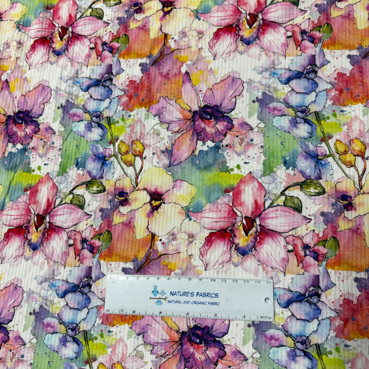 Vibrant watercolor floral print fabric by Nature's Fabrics, featuring pink, purple, and blue blossoms with green leaves on natural organic material.