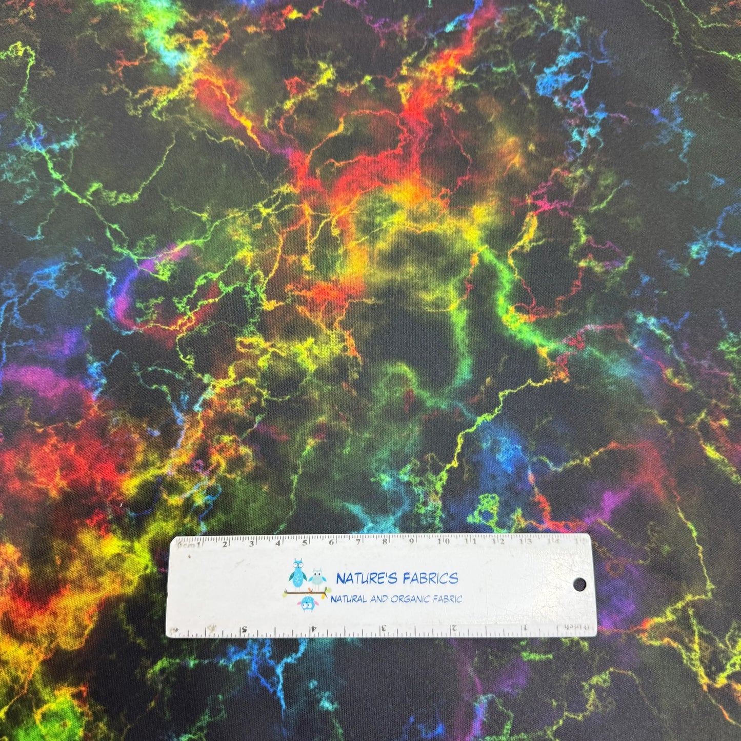 Vibrant neon tie-dye fabric by Nature's Fabrics, featuring rainbow swirls on dark background with measurement ruler.