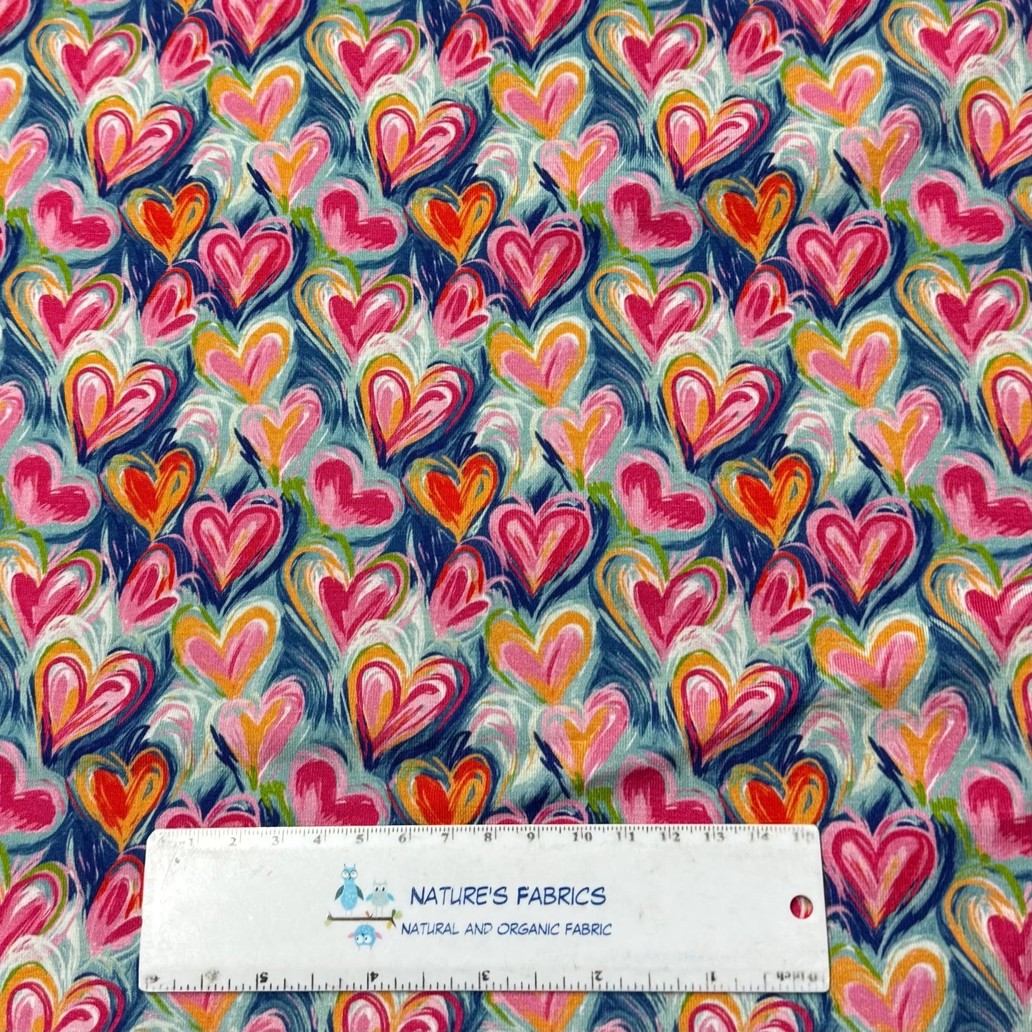 Colorful heart print organic cotton fabric by Nature's Fabrics, featuring pink, red, and orange hearts on a blue background with ruler for scale.