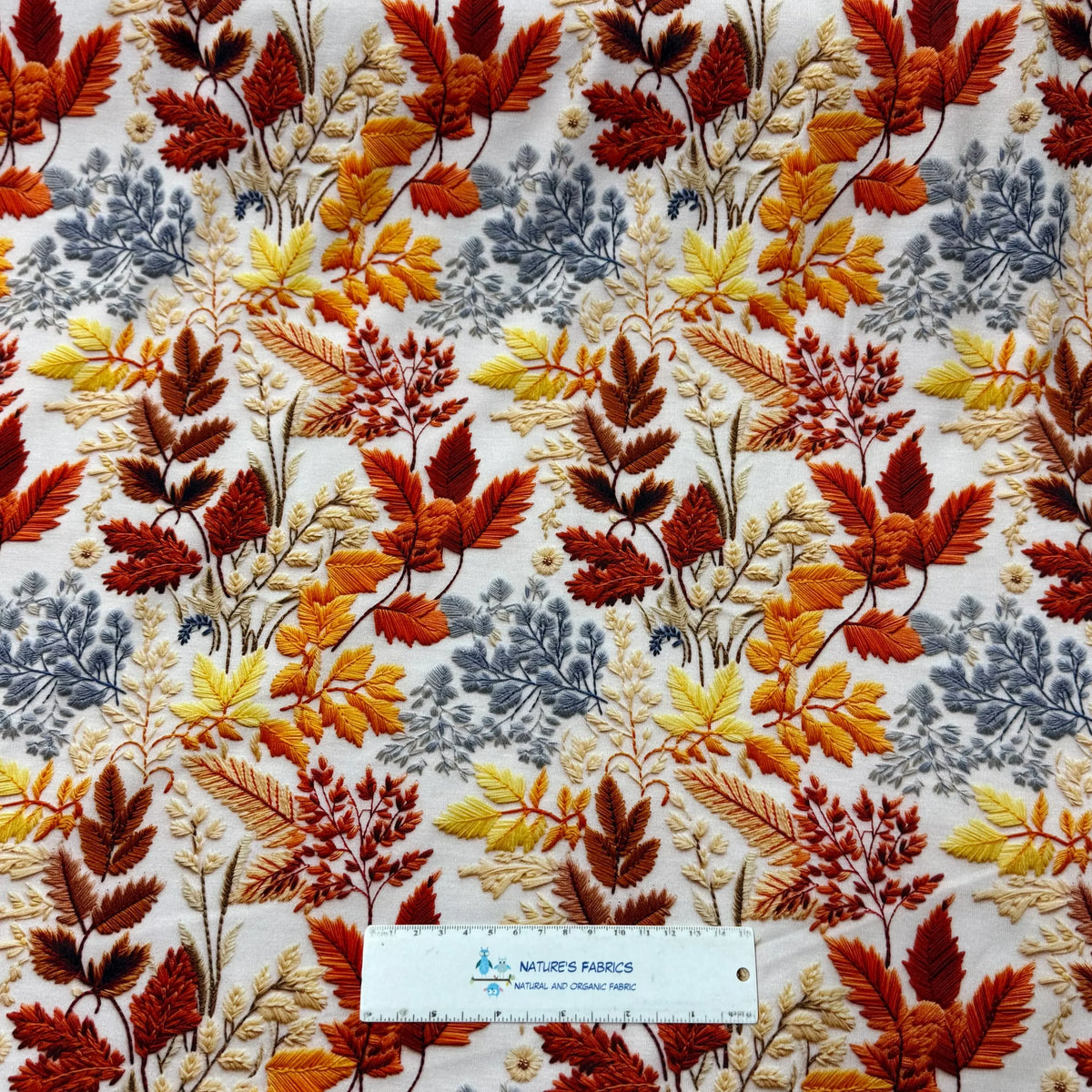 Nature's Fabrics embroidered autumn leaf pattern fabric in orange, gold, and grey tones.