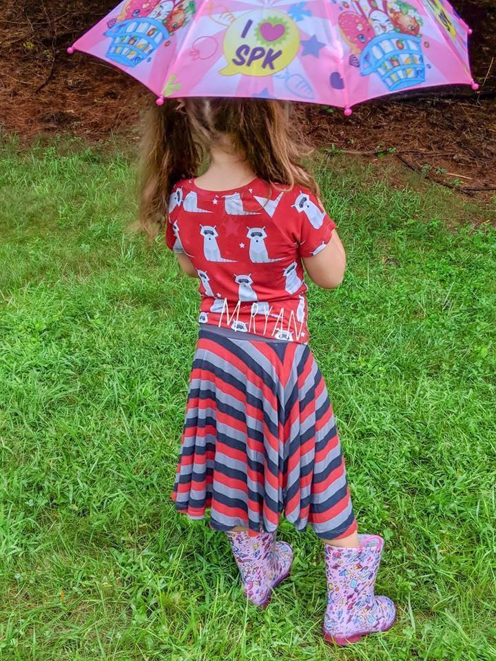 Young girl in Nature's Fabrics red panda shirt and striped skirt holding a colorful umbrella.