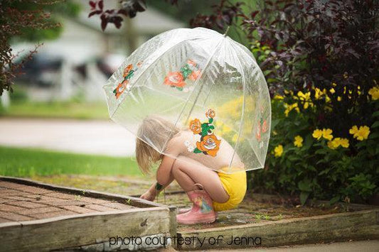 A young child wearing yellow shorts and pink boots under a Nature's Fabrics clear umbrella.
