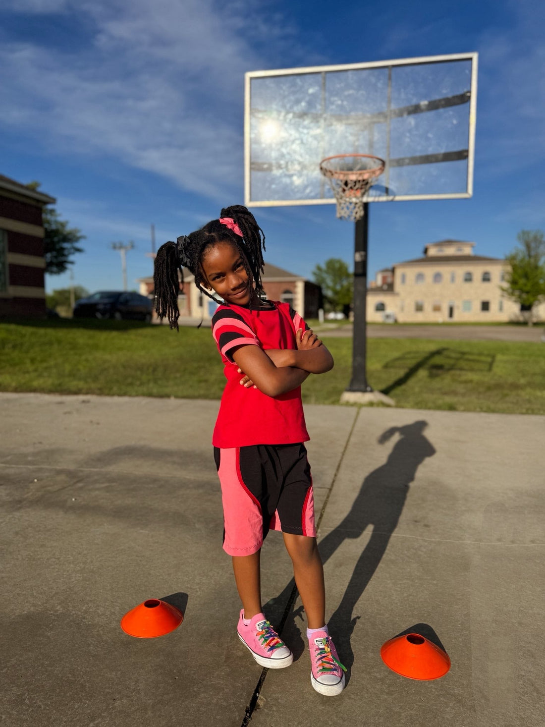 Smiling girl in athletic clothes poses on outdoor basketball court with cones and hoop.